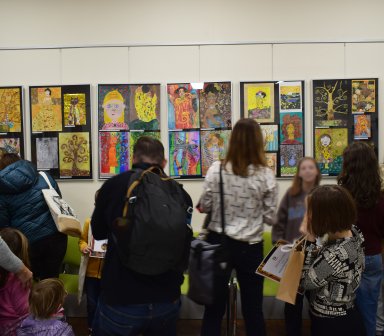 Ludzie widoczni tyłem stojący przed powieszonymi na ścianie pracami plastycznymi umieszczonymi w antyramy. Prace są kolorowe i różnorodne.