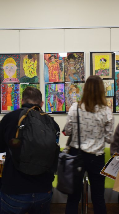 Ludzie widoczni tyłem stojący przed powieszonymi na ścianie pracami plastycznymi umieszczonymi w antyramy. Prace są kolorowe i różnorodne.