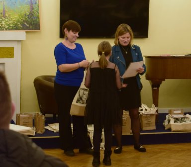 Widzimy organizatorki konkursu, przekazujące jednej z laureatek nagrodę oraz dyplom. W tle przyjemny obrazek łąki, ekran oraz fortepian. Klimat miejsca jest ciepły.