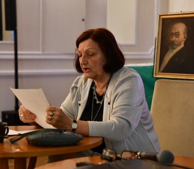 Prelegentka czyta z kartki podczas wykładu. Za nią na stoliku stoi obraz mężczyzny (postać istotna dla wydarzenia Dni esperanto.