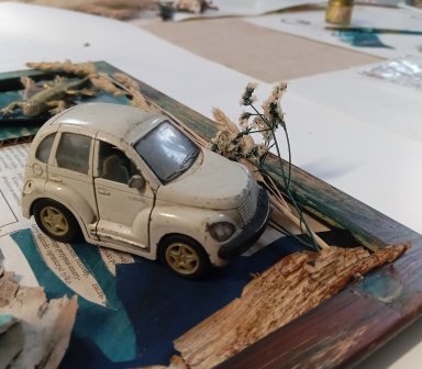 Zdjęcie wykonane pod kątem, skupiające się na detalu białego zabawkowego samochodu (model przypominający Chryslera PT Cruisera), który jest przytwierdzony do wnętrza ramy. Samochód jest zakurzony i nosi ślady użytkowania. Obok niego znajdują się suche kło