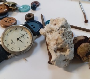 Zbliżenie na grupę przedmiotów: klasyczny zegarek naręczny marki „Perfect” na czarnym pasku, duży kawałek jasnego pumeksu lub porowatego kamienia, zasuszony owoc (orzech), kilka rdzawych gwoździ oraz różnokolorowe guziki porozrzucane na białym blacie.