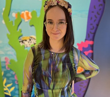 Osoba dorosła, kobieta, w kostiumie inspirowanym motywami podwodnymi, stojąca na tle kolorowej scenografii ekspozycji.