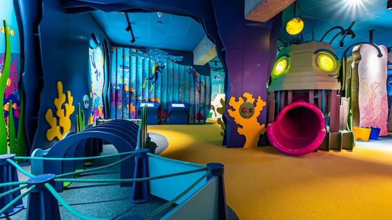 Kolorowa sala dla dzieci z dużą konstrukcją do zabawy, zjeżdżalnią, miękkimi platformami i motywami morskimi.