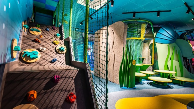 Przestrzeń edukacyjno-rekreacyjna z elementami wspinaczkowymi, siatkami zabezpieczającymi i miękką nawierzchnią w różnych kolorach.