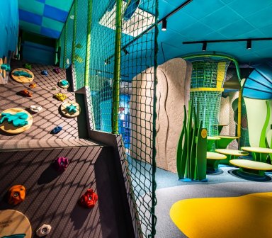 Przestrzeń edukacyjno-rekreacyjna z elementami wspinaczkowymi, siatkami zabezpieczającymi i miękką nawierzchnią w różnych kolorach.