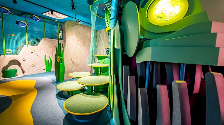 Kolorowa sala edukacyjno-zabawowa inspirowana podwodnym światem, z ilustracjami ryb na ścianach, interaktywnymi ekranami i miękkimi elementami do zabawy.