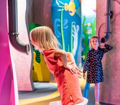 Dziecko pochylone przy instalacji edukacyjnej, mówiące do tuby dźwiękowej oraz drugie, słuchające dźwięków przez tubę umieszczoną w ścianie ekspozycji.