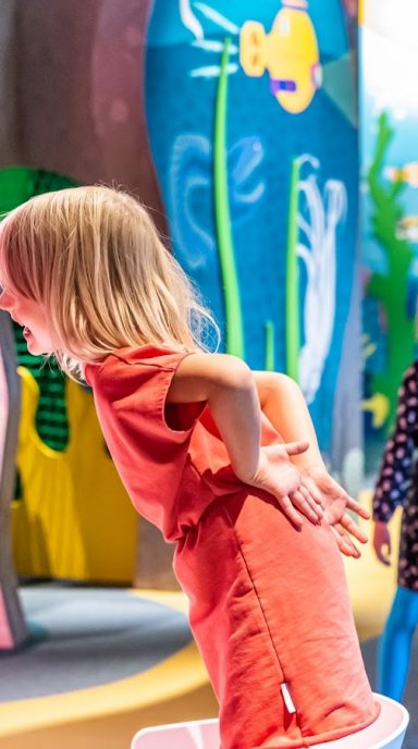 Dziecko pochylone przy instalacji edukacyjnej, mówiące do tuby dźwiękowej oraz drugie, słuchające dźwięków przez tubę umieszczoną w ścianie ekspozycji.