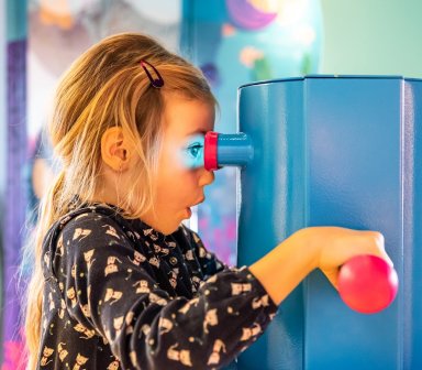 Dziecko korzystające z interaktywnego stanowiska edukacyjnego, zaglądające przez wizjer i obracające elementem urządzenia.