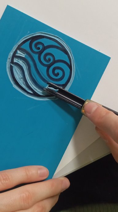 Zbliżenie na dłonie grawerujące dłutem okrągły wzór z motywem fal na niebieskiej matrycy do linorytu.