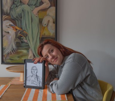 Małgorzata Stasiak siedząca przy stole prezentując swój obraz w ramce.