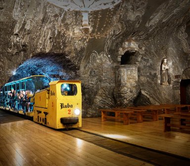 Fot. Kopalnia Soli "Bochnia". Żółta kolejka z wagonikami pełnymi turystów jedzie przez oświetlony, wykuty w soli tunel. W ścianach widoczne są rzeźby i kapliczne zdobienia z soli.