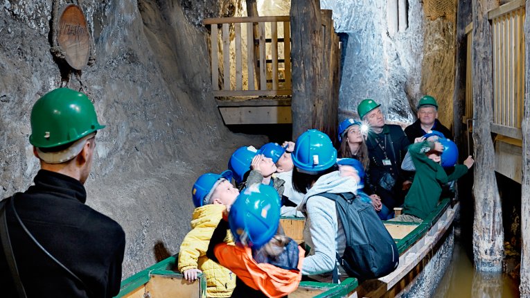 Fot. Kopalnia Soli "Bochnia". Grupa dzieci i dorosłych w kaskach ochronnych płynie drewnianą łodzią przez wąski solny korytarz. Po obu stronach ciągną się wysokie, jasne skalne ściany, a nad nimi drewniane pomosty.