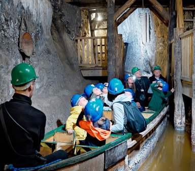 Fot. Kopalnia Soli "Bochnia". Grupa dzieci i dorosłych w kaskach ochronnych płynie drewnianą łodzią przez wąski solny korytarz. Po obu stronach ciągną się wysokie, jasne skalne ściany, a nad nimi drewniane pomosty.