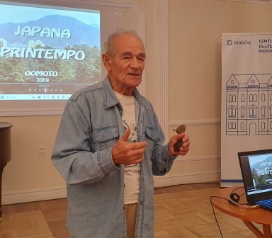Mówca prowadzi wykład, a za jego plecami wyświetlany na rzutniku jest obraz przyrodniczy z napisami w języku esperanto.
