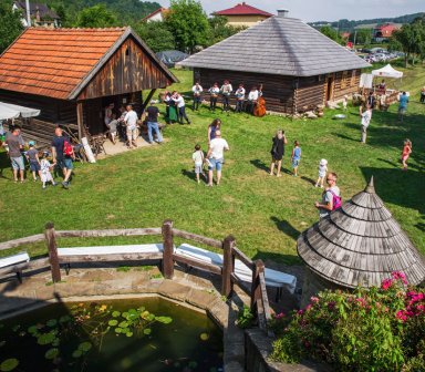 Fot. Zagroda w Jędrzejkówce. Widok z góry na skansenowy plac z drewnianymi chatami. Na trawie spacerują rodziny z dziećmi, a obok jednej z chat gra zespół ludowy. W pierwszym planie widać niewielki staw z nenufarami i drewniany mostek.