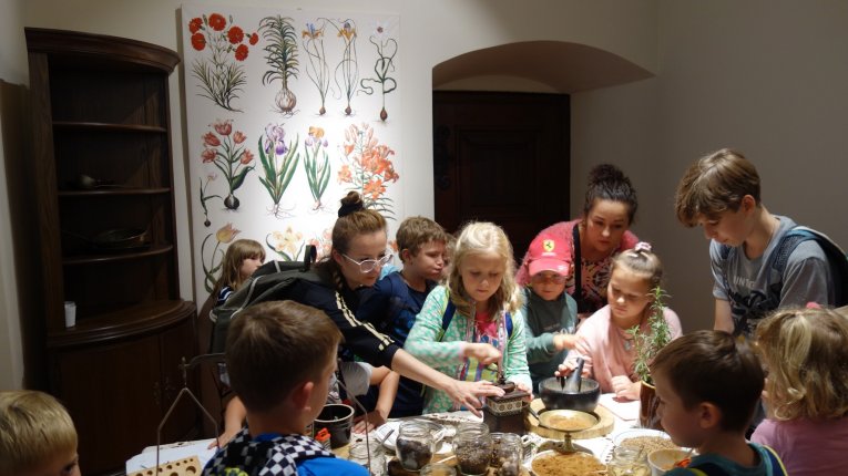 Grupa dzieci wraz z opiekunami uczestniczy w warsztatach edukacyjnych przy stole, przygotowując mieszanki ziół i nasion; na stole stoją słoiki z roślinami i moździerze, w tle plansza z ilustracjami botanicznymi.