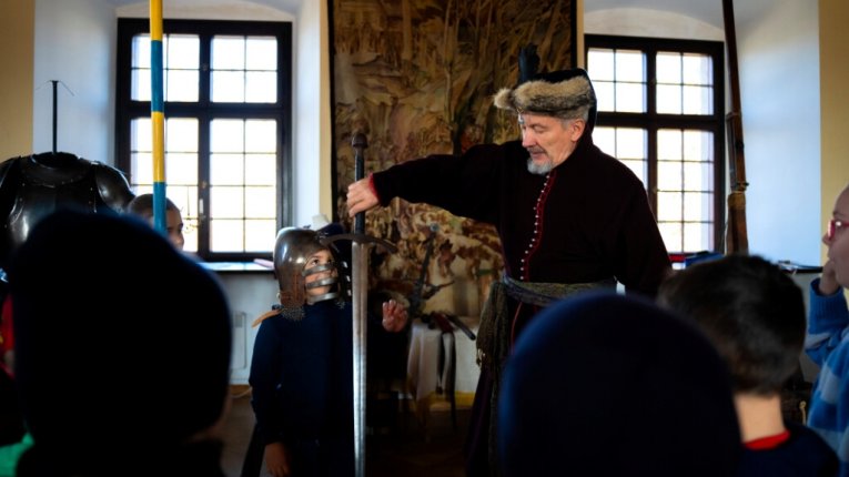 Fot. Zamek w Wiśniczu. Mężczyzna w historycznym stroju pokazuje dzieciom długi miecz. Jedno z dzieci ma na głowie ciężki rycerski hełm. W tle widać duże okna i średniowieczną zbroję.