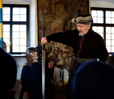 Fot. Zamek w Wiśniczu. Mężczyzna w historycznym stroju pokazuje dzieciom długi miecz. Jedno z dzieci ma na głowie ciężki rycerski hełm. W tle widać duże okna i średniowieczną zbroję.