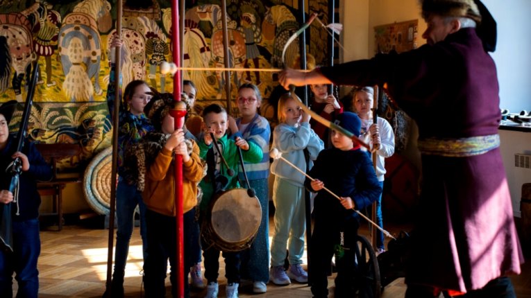 Fot. Zamek w Wiśniczu. Dzieci stoją w półkolu, każde trzyma historyczną broń lub rekwizyt. Instruktor w stroju z epoki napina łuk, pokazując, jak strzelać. W tle kolorowa tkanina z dawnymi wzorami.