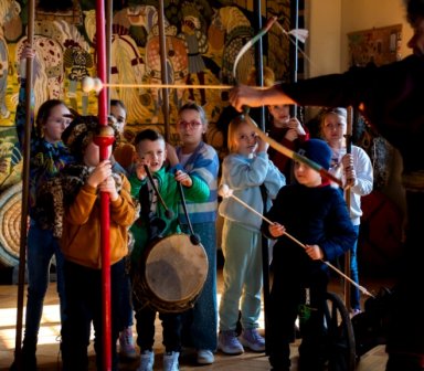 Fot. Zamek w Wiśniczu. Dzieci stoją w półkolu, każde trzyma historyczną broń lub rekwizyt. Instruktor w stroju z epoki napina łuk, pokazując, jak strzelać. W tle kolorowa tkanina z dawnymi wzorami.