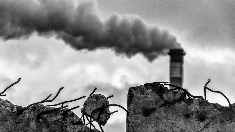 Zbliżenie na spękaną, krawędź betonowego muru, z której wystają wygięte, ciemne druty zbrojeniowe. W tle, na podchmurnym niebie, widoczny jest wysoki przemysłowy komin, z którego unosi się gęsty, ciemny dym.